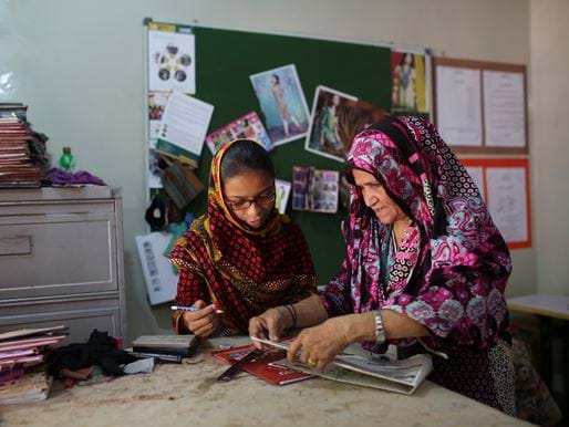 Woman teaching sewing design skills to a younger woman in front of pin board, Pakistan